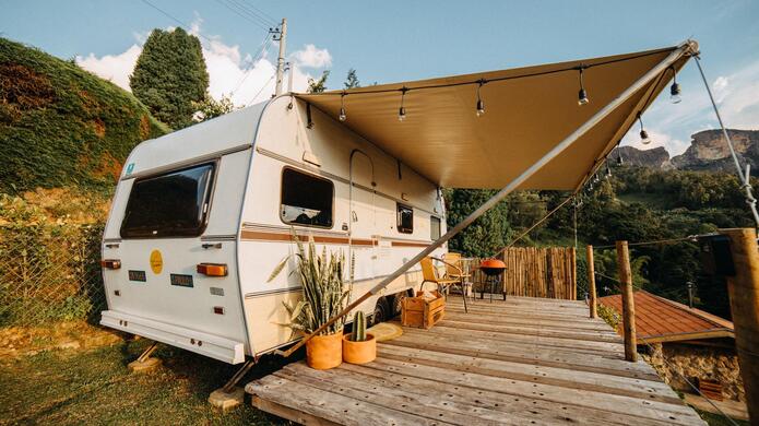 Camper con tendalino esteso su una terrazza in legno immersa in un paesaggio verde con alberi e montagne sullo sfondo.
