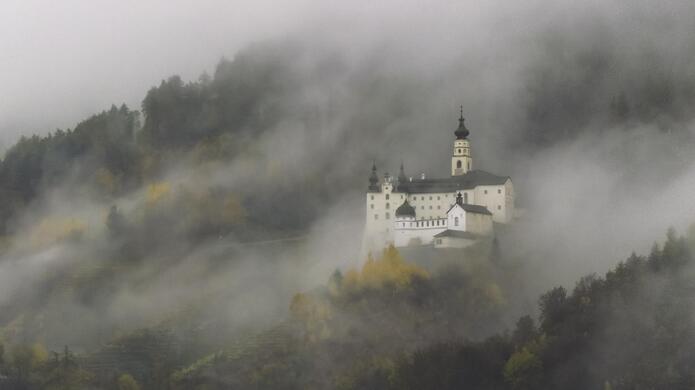 Kloster Marienberg umgeben von bewaldeter Hügellandschaft im Nebel.