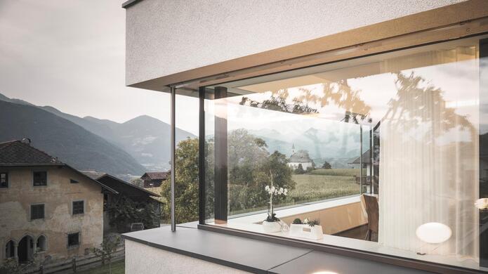 Große Fensterfront mit Blick in einen Wohnraum, die umliegenden Häuser und grüne Landschaft.
