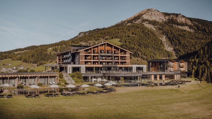 Hotelgebäude Granvara in alpiner Landschaft mit großer Terrasse, umgeben von einer grünen Wiese und Bergen im Hintergrund bei bewölktem Himmel.