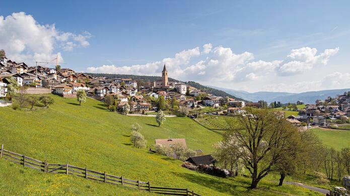 Blick auf das Dorf Jenesien mit grünen Wiesen im Vordergrund und blauem Himmel im Hintergrund.